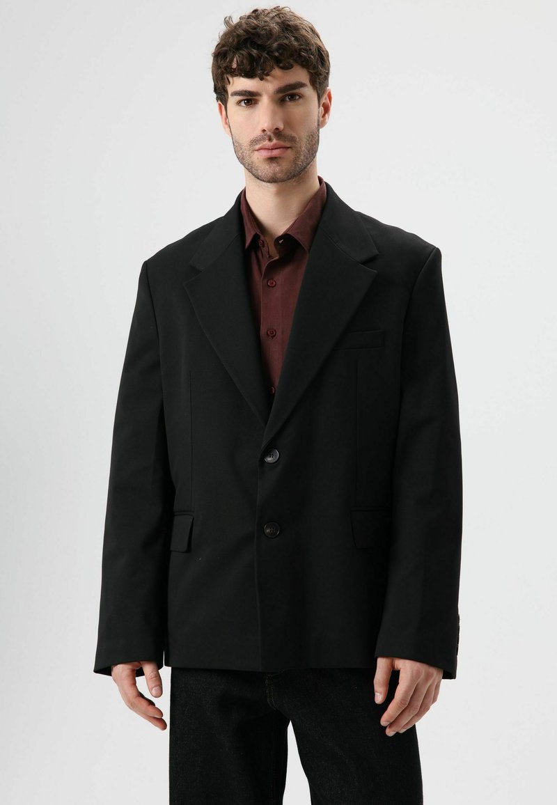 Zwarte blazer met een ingesneden revers, twee knopen en twee steekzakken aan de voorkant, gedragen over een bordeauxrode blouse, met een gladde stoftextuur.