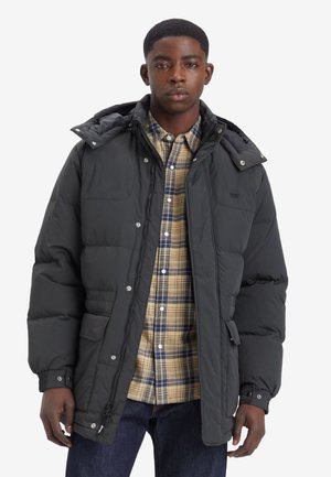 Padded black jacket with a hood, zip closure, and snap buttons. Worn over a beige and blue checkered shirt, displayed against a white background.