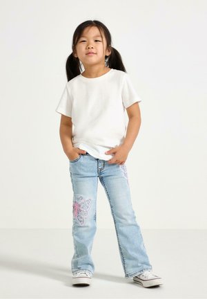 Bambino che indossa una maglietta bianca a maniche corte e jeans azzurri svasati con ricamo di farfalla rosa sul davanti. Scarpe da ginnastica bianche.