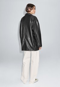 Zwarte leren jas met een ontspannen pasvorm, voorzien van een sjaalkraag en zichtbare naaddetails, gecombineerd met lichtgekleurde broek met wijde pijpen.