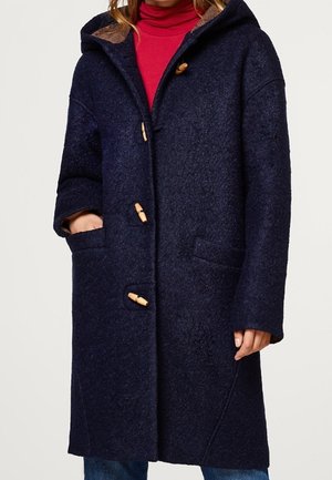 Woman wearing a navy blue hooded duffle coat with wooden toggle fastenings and red turtleneck underneath, hands in coat pockets.