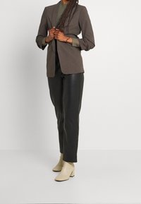 Blazer marrone su misura, tessuto texturizzato, frontale aperto, abbinato a pantaloni neri slim-fit e stivaletti alla caviglia beige chiaro.
