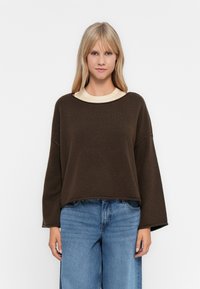 Suéter castanho, oversized, com decote arredondado e ombros caídos. Usado sobre uma t-shirt creme, acompanhado de jeans denim azul claro.