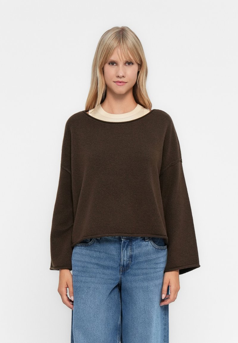 Suéter castanho, oversized, com decote arredondado e ombros caídos. Usado sobre uma t-shirt creme, acompanhado de jeans denim azul claro.