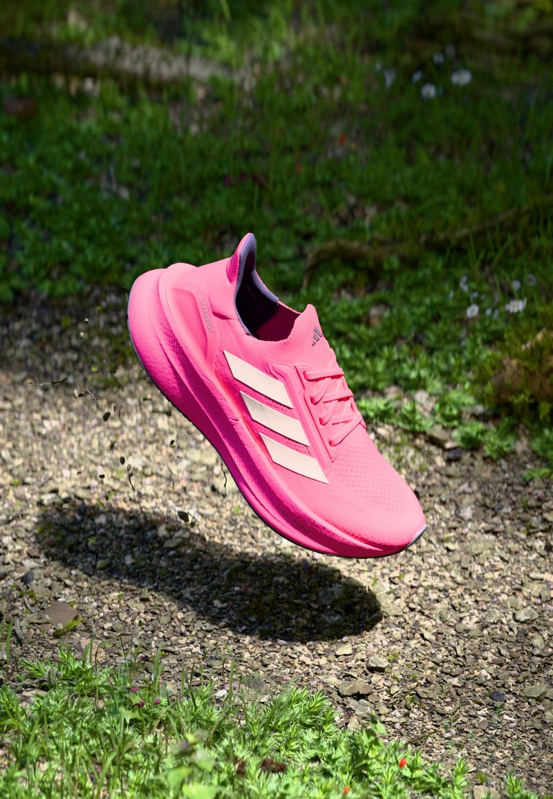 Shoe de corrida Adidas rosa brilhante no ar acima de uma caminho de cascalho, com relva verde e pequenas flores ao fundo.