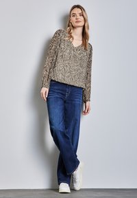Leichte, gemusterte Bluse in gedämpftem Olivgrün, mit V-Ausschnitt und langen Ärmeln; kombiniert mit dunkelblauen, weiten Jeans und weißen Sneakers.
