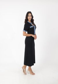 Zwart geribbelde midi-jurk met korte mouwen en denim accenten. Voorzien van een knoopsluiting aan de voorkant en twee borstzakken. Transparante blokhak sandalen.