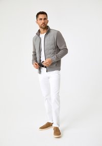 Graue gepolsterte Jacke mit hohem Kragen, weißes T-Shirt, weiße Hose und braune Slipper. Strukturmaterial und Reißverschlussdetails sind sichtbar.