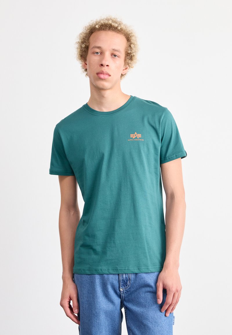 Zelena bombažna majica s kratkimi rokavi in krožnim izrezom, z majhnim oranžnim grafičnim logotipom na levi strani prsnega koša. Sproščen kroj.