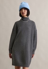 Grijze gebreide sweater jurk met een hoge kraag, verlaagde schouders en geribbelde manchetten. Gepaard met een lichtblauwe gebreide muts. Zilveren kettingdetail.