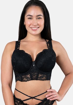 Frau lächelt, trägt ein schwarzes Spitzen-Bralette und passende, geschnürte Spitzenunterwäsche, posiert mit Hand in der Hüfte vor einem schlichten Hintergrund.