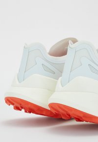 Buty sportowe z białą cholewką, jasnoniebieskimi akcentami i teksturowanym obcasem. Wyróżniają się kontrastującą pomarańczową podeszwą z gumy, zapewniającą przyczepność.