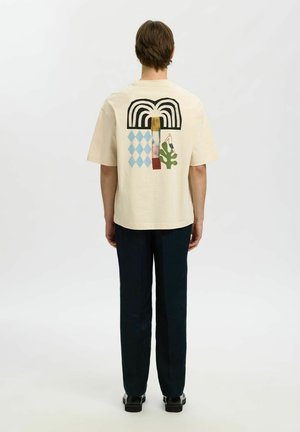 Mann steht mit dem Rücken zur Kamera, trägt ein beiges T-Shirt mit abstrakter Kunst, schwarze Hose und schwarze Schuhe vor weißem Hintergrund.
