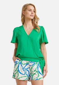 Grünes Wickeltop mit kurzen Ärmeln und strukturiertem Finish. Kombiniert mit weißen Shorts, die mit grünen und blauen Blattmustern verziert sind.