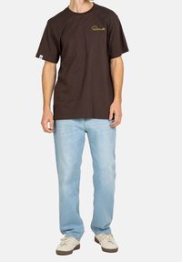 Camiseta de algodón marrón con mangas cortas, con un logotipo amarillo en el pecho, combinada con vaqueros azul claro y zapatillas beige.