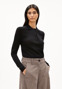 Frau mit langen, dunklen Haaren trägt ein schwarzes, knopfbares Langarmhemd, das in hochgeschnittene, braune Textilhosen gesteckt ist, mit den Händen in den Taschen.