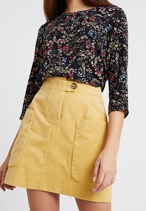 Frau trägt eine schwarze Bluse mit Blumenmuster und drei Viertelärmeln sowie einen gelben Cordrock mit großen Fronttaschen.