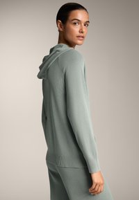 Svetlozeleni rebrasti dukserica s kapuljačom na zavlačenje, dugim rukavima i teksturiranim materijalom. Prikazana iz bočnog ugla kako bi se naglasile detalje.