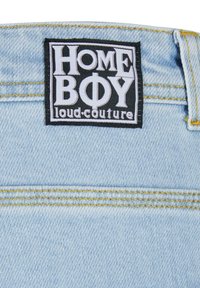 Gaiši zils džinsu audums ar redzamām šuvēm. Melna etiķete jostā ar uzrakstiem "HOME BOY" un "loud-couture" baltā krāsā.