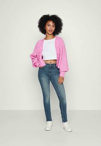Cardigan de canalé rosa, top corto blanco y vaqueros ajustados azules. El cardigan tiene botones y mangas voluminosas, mientras que los vaqueros tienen un lavado desvanecido.