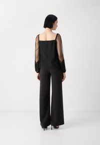 Schwarzes Oberteil mit durchsichtigen langen Ärmeln und Spitze, kombiniert mit weiten schwarzen Hosen und hohen Schuhen. Quadratischer Ausschnitt, glatte Textur.