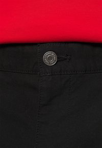 Pantalones negros con cierre de botón en la parte delantera, cordón y textura de tela suave, mostrados parcialmente sobre un fondo rojo.