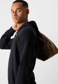 Junger Mann in einem schwarzen Hoodie, der eine braune Ledertasche über der Schulter trägt und über die Schulter schaut, vor einem schlichten Hintergrund.
