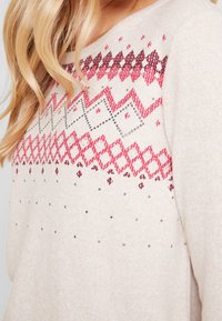 Maglione beige a maglia con motivi geometrici in rosso e rosa scuro. Presenta piccoli borchiette argentati e un collo a coste, offrendo un aspetto testurizzato.