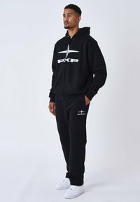 Schwarze Zip-Hoodie mit weißem "PXP"-Logo, kombiniert mit passenden Jogginghosen mit einem weißen Logo-Akzent. Getragen mit weißen Sneakers.