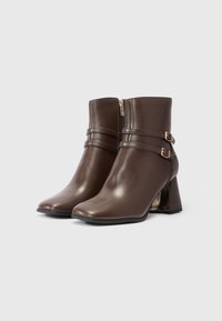 Botas de cuero marrón hasta el tobillo con tacones de bloque, cremalleras laterales y correas con doble hebilla, exhibidas sobre un fondo blanco liso.