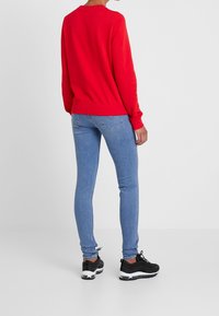 Röd sweatshirt med rund halsringning och långa ärmar, i kombination med ljusblå skinny jeans och svarta sportskor. Bakifrån.