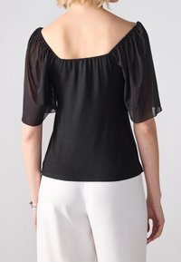 Černá blůza s vyklenutými průhlednými rukávy do loktů, nařaseným výstřihem a těsným střihem; nošena s bílými kalhotami s vysokým pasem.