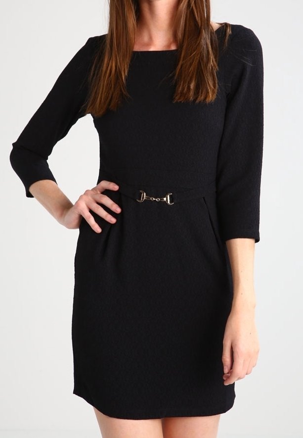 Femme portant une robe noire longueur genou avec des manches trois-quarts et une petite ceinture à boucle métallique à la taille, sur un fond uni.