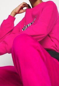 Sudadera rosa con un logo a rayas de cebra en blanco y negro, combinada con pantalones de chándal rosa a juego. Tejido suave y texturizado, corte holgado.