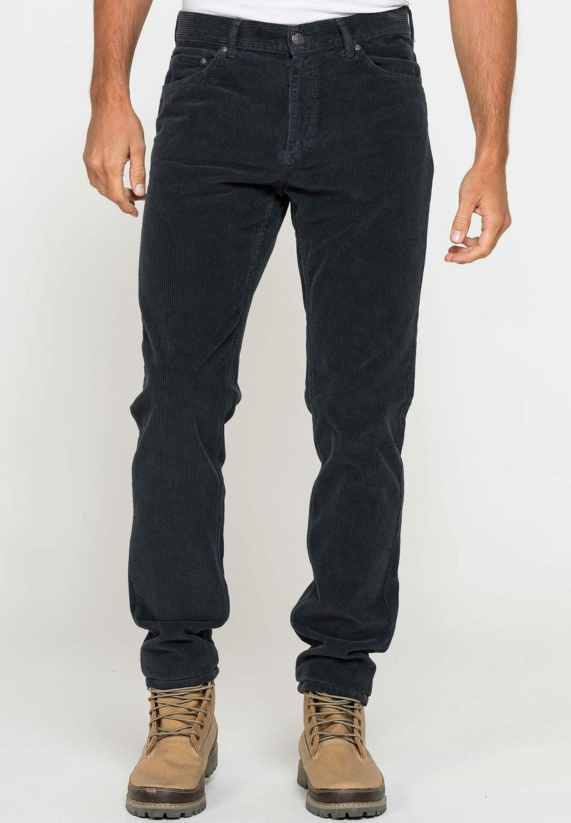 Pantaloni di velluto a coste scuri con vestibilità slim, dotati di zip frontale, tasche anteriori e una evidente texture a coste verticali. Indossati con stivali beige.