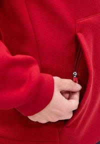 Felpa rossa realizzata in tessuto morbido, con una tasca laterale con zip, cerniera liscia e polsini aderenti. Dettaglio notevole include il tirazip.