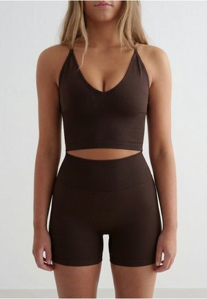 Kvinne iført en tettsittende mørk brun crop top med tynne stropper og matchende høytlivs biker shorts mot en enkel lys bakgrunn.