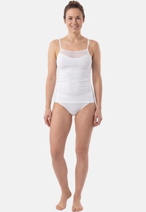 Donna sorridente in piedi a piedi nudi che indossa una canottiera bianca con pizzo e un intimo bianco in stile bikini abbinato, su sfondo neutro.