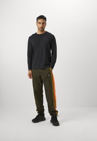 Langärmliges schwarzes Hemd mit dezentem Logo, kombiniert mit olivgrünen Hosen mit orangefarbenen Seitenstreifen und einem Logo-Patch. Schwarze Schuhe.
