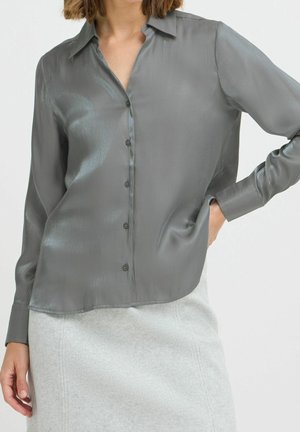 Vrouw die een glanzende grijze blouse met knopen en lange mouwen draagt en een lichtgrijze rok, staand met één hand in haar zak.