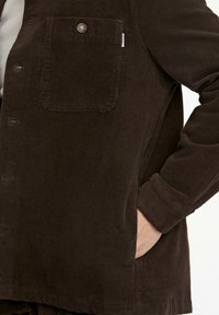 Brun kordflodsskjorta med knäppning, en bröstficka och ribbad textur, med en avslappnad passform och uppvikt ärm.