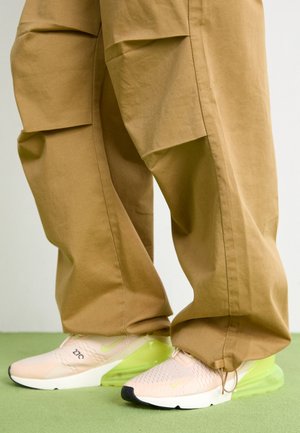 Lichtroze en limoengroene sportschoenen gedragen met losvallende bruine cargobroeken, staand op een groene vloer.