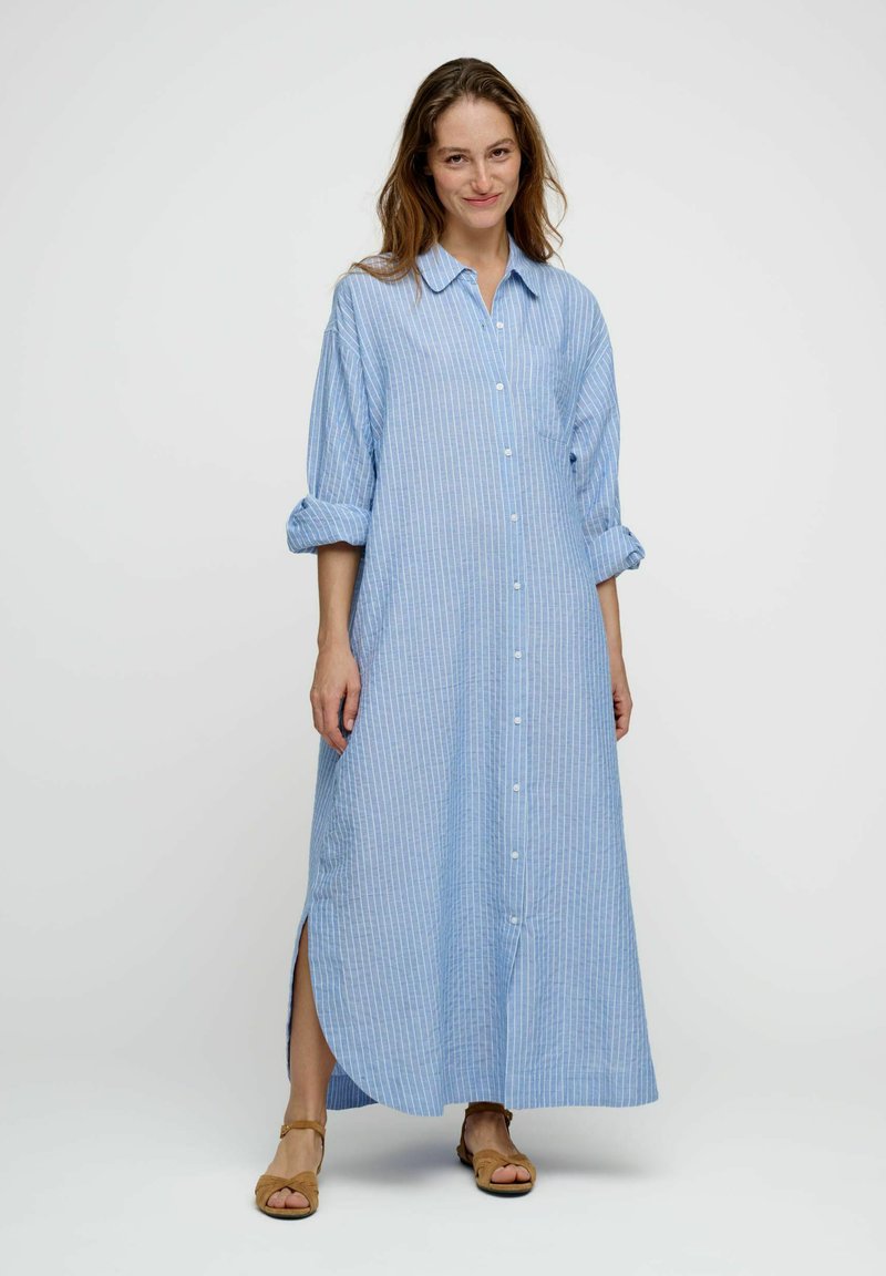 Femme portant une longue robe chemise à rayures bleues et blanches avec manches retroussées et sandales beiges, debout devant un fond uni.