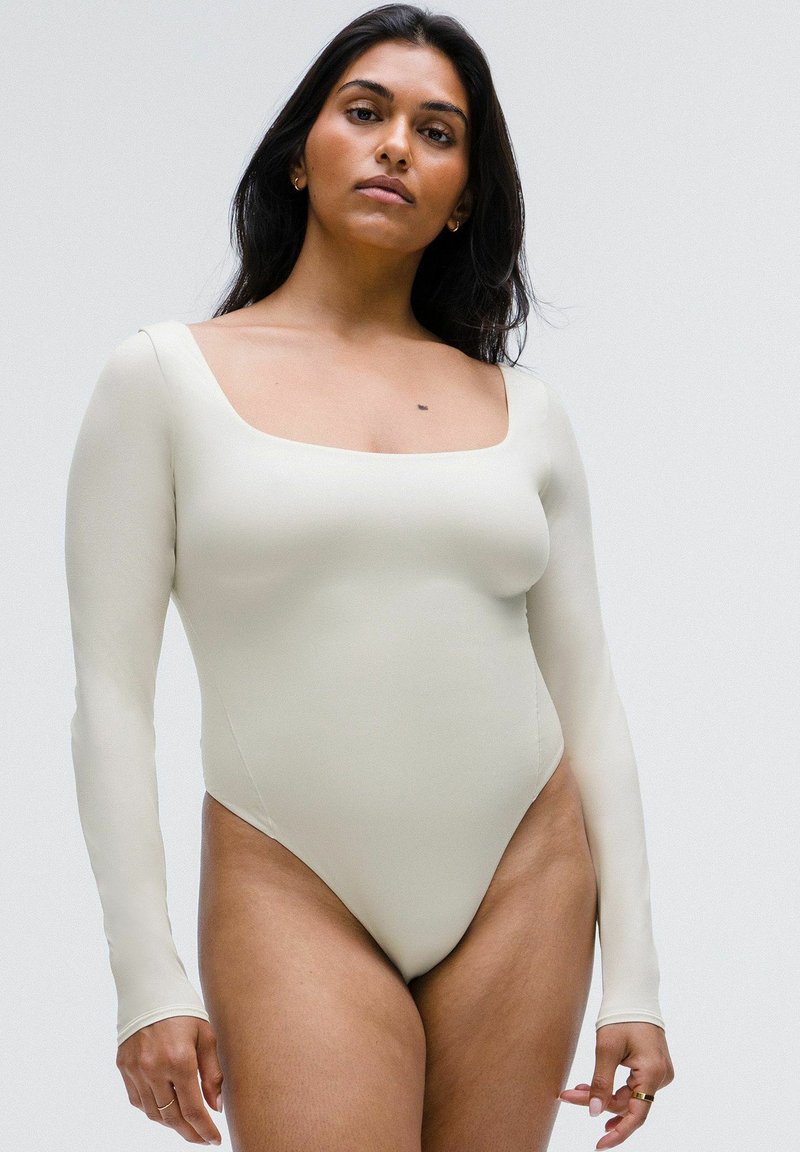 Bodysuit de manga larga en suave tejido beige con escote cuadrado. Textura suave, diseño ajustado y detalles de costura mínimos.