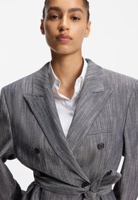 Blazer rayé gris à double boutonnage, boutons ton sur ton et taille ceinturée, associé à une chemise blanche à col en dessous.