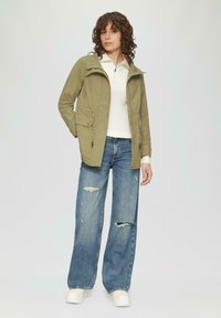 Giacca leggera verde oliva con vita regolabile e due tasche frontali, abbinata a jeans a gamba larga blu chiaro con effetti usurati e sneakers bianche.