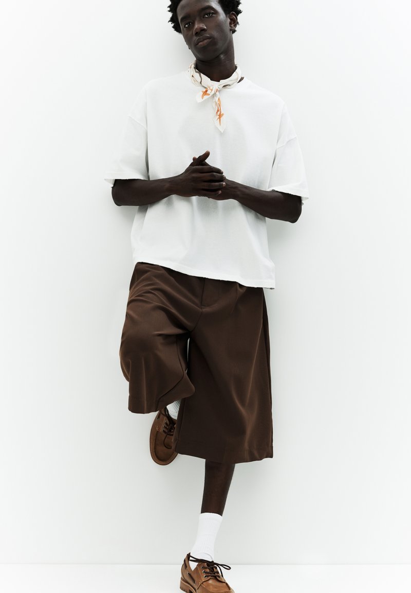 Hvid oversized t-shirt, brune vide shorts, brune sko og hvide sokker. Tilbehør: orange og beige mønstret tørklæde om halsen.