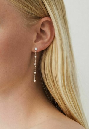 Persoană blondă purtând o cercei lungi de argint cu cinci perle mici albe, prim-plan al urechii și părului pe un fundal neutru.