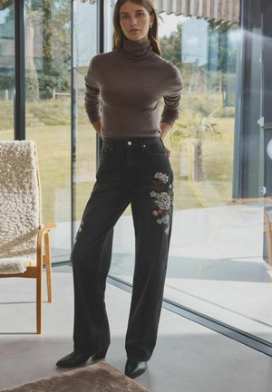 Schwarze weit geschnittene Jeans mit floralem Stickerei am linken Bein, kombiniert mit einem taillierten braunen Rollkragenpullover und schwarzen High Heels Stiefeletten.