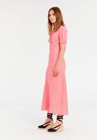 Robe rose mi-longue avec des manches évasées et un détail nœud au niveau du cou. Portée avec des chaussures compensées en espadrilles noires et beiges.